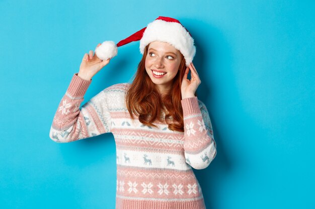 Conceito de inverno e véspera de Natal. Linda garota adolescente com camisola e chapéu de Papai Noel, comemorando o Natal, sorrindo feliz, em pé sobre um fundo azul.