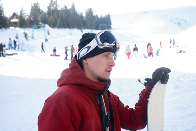 Conceito de inverno, lazer, esporte e pessoas - plano aproximado de um snowboarder em pé em sua prancha com vista para uma pista de esqui na encosta nevada em um resort de inverno
