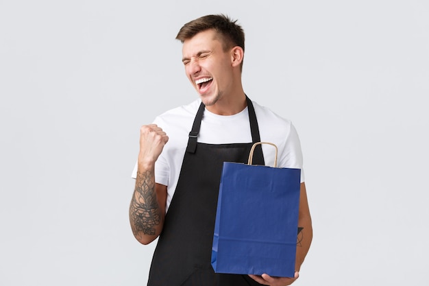 Foto grátis conceito de loja, compras e funcionários de varejo. vendedor alegre e feliz, finalmente trabalhando após covid-19, balançando o punho e cantando de felicidade, segurando uma bolsa ecológica com fundo branco