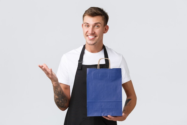 Conceito de loja, compras e funcionários de varejo. vendedor louro e sorridente carismático na loja, conversando com o cliente, embalando produtos em eco-bolsas, fundo branco de pé com aparência amigável