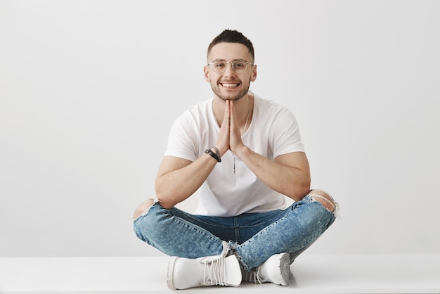 Conceito de meditação e emoções Atraente elegante jovem macho com barba usando óculos e roupa da moda sentado com as mãos cruzadas no chão enquanto de mãos dadas em oração e sorrindo sobre parede cinza