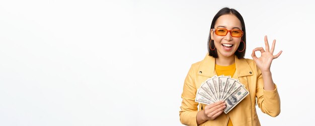 Conceito de microcrédito e empréstimos, garota coreana elegante e feliz, mostrando sinal de ok e dinheiro em dinheiro