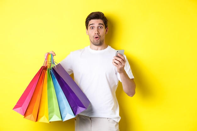 Conceito de mobile banking e cashback. homem surpreso, segurando sacolas de compras e smartphone, em pé sobre um fundo amarelo.