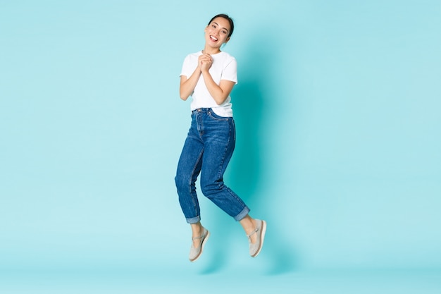 Conceito de moda, beleza e estilo de vida. Feliz e sonhadora, excitada garota asiática em roupa casual, pulando de felicidade e alegria, batendo palmas com entusiasmo, em pé sobre a parede azul claro