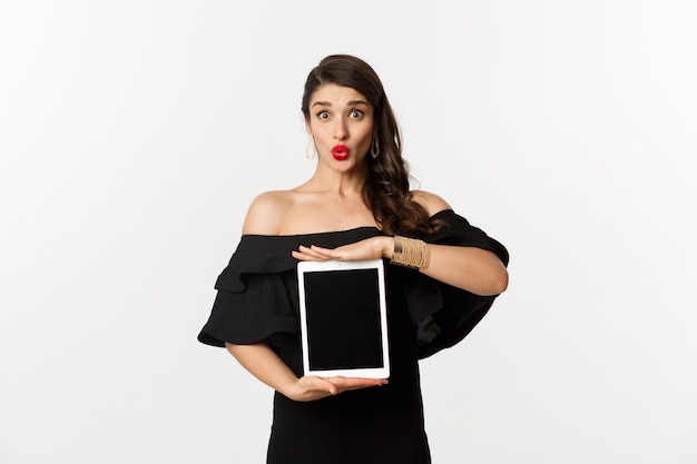 Conceito de moda e compras. Mulher bonita com batons vermelhos, vestido preto, mostrando a tela do tablet e parecendo animada, em pé sobre um fundo branco.