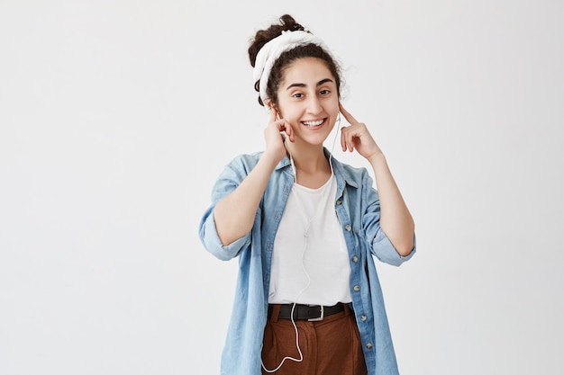 Conceito de música e tecnologia. Menina de cabelos escuros ouvindo audiobook ou rádio no celular com fones de ouvido, olhando e sorrindo contra a parede do espaço branco cópia para conteúdo de publicidade