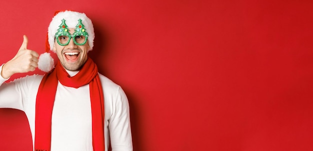 Conceito de natal, férias de inverno e celebração. Cara alegre de chapéu de Papai Noel e óculos de festa, mostrando o polegar para cima e sorrindo satisfeito, de pé sobre fundo vermelho