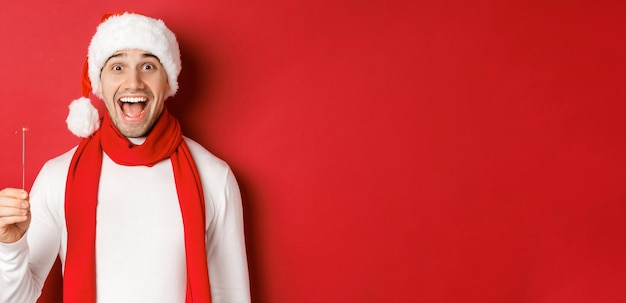 Foto grátis conceito de natal, férias de inverno e celebração. close-up de feliz homem bonito com chapéu de papai noel e lenço, se divertindo na festa de ano novo, segurando o diamante e sorrindo, fundo vermelho.