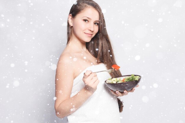 Conceito de neve, inverno, natal, beleza, cuidados com a pele e pessoas - linda garota na toalha, comendo salada fresca e sorrindo. mulher comendo legumes frescos na cozinha sobre fundo de neve Foto Premium