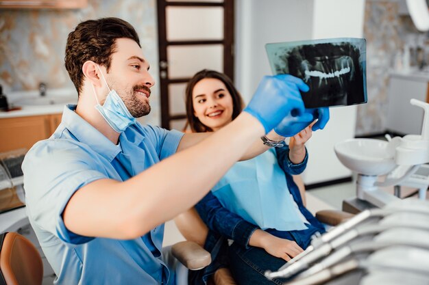 Conceito de odontologia e saúde, dentista masculino mostrando raio-x de dentes para paciente do sexo feminino na sala de clínica odontológica branca.