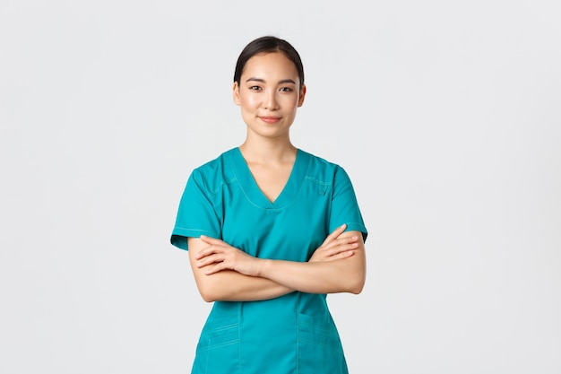 Conceito de pandemia de trabalhadores de saúde covid19 Enfermeira asiática sorridente confiante em jaleco em pé peito de braços cruzados confiante Médico profissional pronto para cirurgia em pé fundo branco