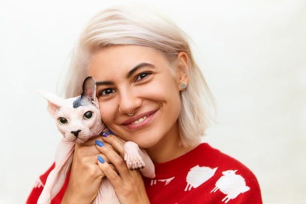 Conceito de pessoas, animais de estimação, animais domésticos, amizade, amor e cuidado. Imagem isolada de uma jovem europeia bonita em um macacão vermelho segurando o gato Sphynx direito, pressionando o adorável animal de estimação contra o peito e sorrindo