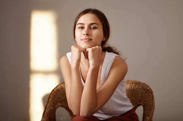 Conceito de pessoas, beleza e juventude. Retrato interior de uma encantadora jovem europeia esbelta bronzeada relaxando em casa, sentada na poltrona tecida sozinha, colocando as mãos sob o queixo. Sol hora dourada