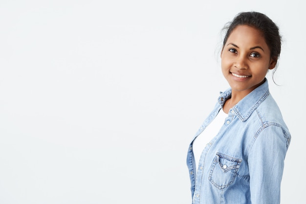 Conceito de pessoas e beleza. Retrato de mulher bonita de pele escura, posando na parede branca com sorriso bonito e charmoso, vestido com camisa jeans azul claro.