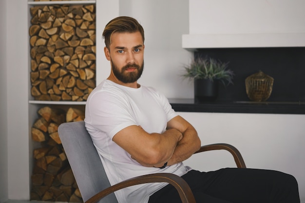 Foto grátis conceito de pessoas e estilo de vida. foto horizontal de um jovem europeu com barba por fazer, corpo atlético e rosto peludo relaxando em casa, mantendo os braços cruzados, com expressão séria