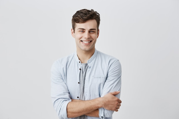 Conceito de pessoas e estilo de vida. homem caucasiano jovem atraente de bom humor, camisa azul de mangas compridas, sorrindo alegremente mostrando os dentes brancos perfeitos, felizes com notícias positivas, mantendo os braços cruzados.