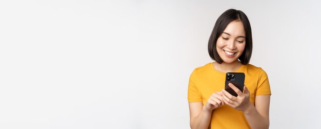 Conceito de pessoas e tecnologia sorrindo menina asiática usando mensagens de smartphone no celular em pé