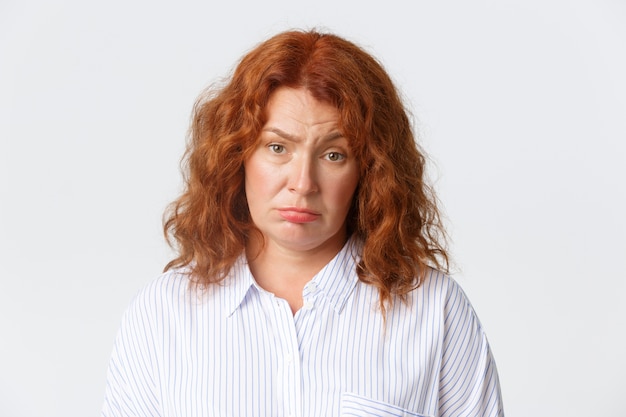 Foto grátis conceito de pessoas, emoções e estilo de vida. mulher de meia-idade ruiva cansada não divertida fazendo cara triste e olhando para a câmera relutante, em pé sombrio sobre fundo branco. copie o espaço