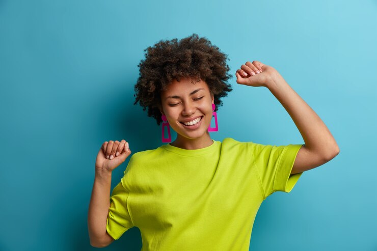 Foto grátis conceito de pessoas, emoções, estilo de vida e lazer. modelo feminino de pele escura, alegre e otimista dança com as mãos para cima, se diverte e festeja, se move ao ritmo da música, tem sorriso alegre, isolado na parede azul