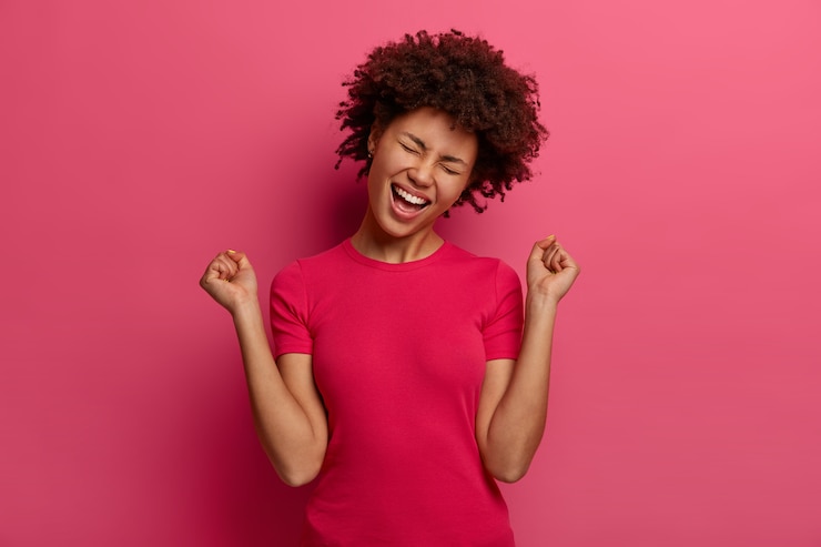 Foto grátis conceito de pessoas, emoções, triunfo e sucesso. feliz adolescente afro-americana comemora vitória, levanta os punhos, tem um humor otimista, vestida com roupas casuais, isolada na parede rosa.