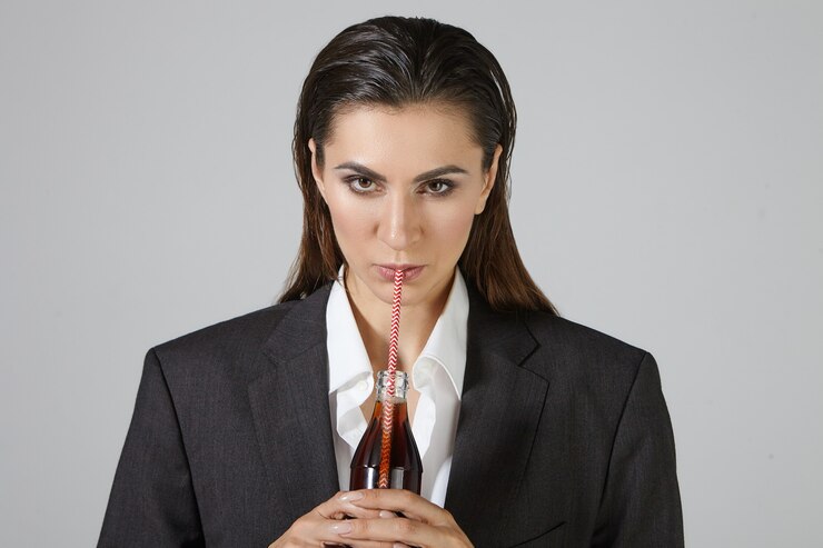 Foto grátis conceito de pessoas, estilo de vida, alimentos e bebidas. foto de uma empresária séria e sedenta, vestida com roupas masculinas enormes, posando isolada segurando uma garrafa de vidro, bebendo refrigerante marrom com canudo