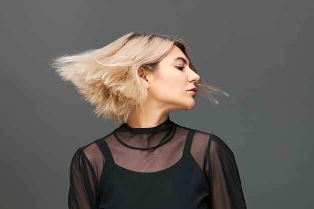Conceito de pessoas, estilo de vida, diversão, festividade e festa. Foto isolada de uma bela jovem encantadora com penteado loiro bob curtindo uma boa música, dançando com os olhos fechados, cabelo esvoaçando ao vento