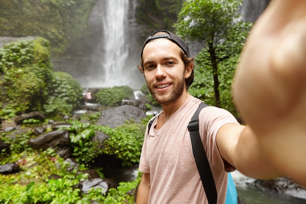 Conceito de pessoas, estilo de vida, natureza e aventura. Elegante jovem viajante com mochila tomando selfie na floresta tropical com cascata