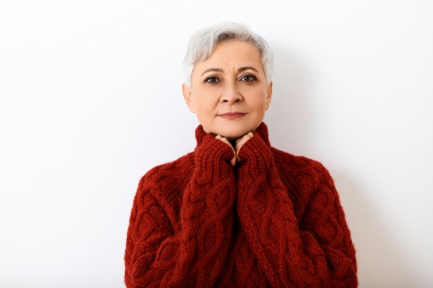 Conceito de pessoas, idade, estilo, moda e temporadas. Foto de uma linda mulher idosa de 60 anos, feliz, com um penteado curto de duende segurando as mãos sob o queixo e sorrindo, vestida com um suéter de tricô