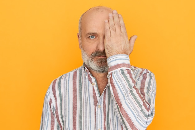 Conceito de pessoas, idade, saúde e aposentadoria. homem aposentado careca com a barba por fazer e camisa listrada cobrindo um dos olhos com a mão, tendo a visão testada em clínica de oftalmologia, não podendo ver objetos próximos