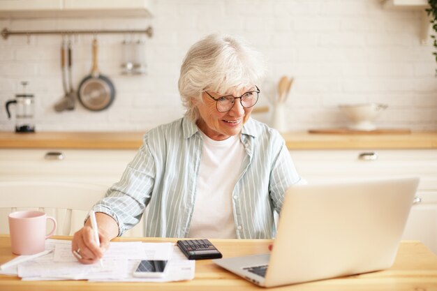Conceito de pessoas, idade, tecnologia e ocupação. imagem interna de uma atraente aposentada sorridente de cabelos grisalhos usando laptop para trabalho remoto, sentada na cozinha com papéis e ganhando dinheiro online
