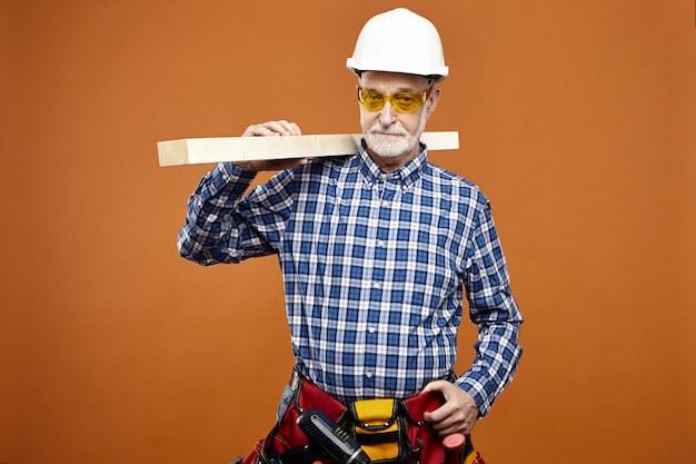 Conceito de pessoas, idade, trabalho e ocupação. Retrato de homem sênior confiante e focado, usando óculos de proteção amarelos, camisa xadrez, capacete e bolsa wiast com ferramentas, carregando uma barra de madeira no ombro