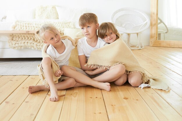 Conceito de pessoas, infância, família, amor e união. Foto doce e aconchegante de três lindos irmãos pequenos sentados no chão de madeira, enrolados em um cobertor, se abraçando