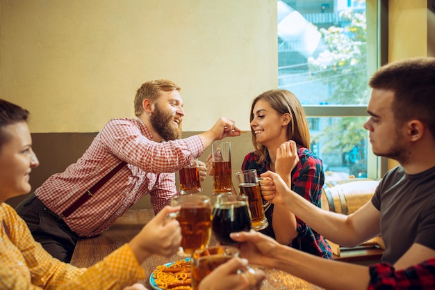 Conceito de pessoas, lazer, amizade e comunicação - amigos felizes bebendo cerveja, conversando e tilintar de copos no bar ou pub