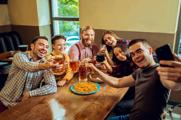 Conceito de pessoas, lazer, amizade e comunicação - amigos felizes bebendo cerveja, conversando e tilintar de copos no bar ou pub