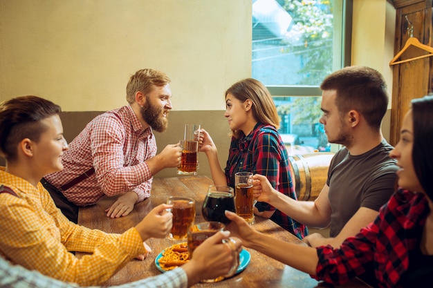 Conceito de pessoas, lazer, amizade e comunicação - amigos felizes bebendo cerveja, conversando e tilintar de copos no bar ou pub