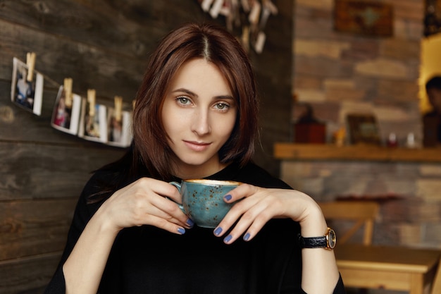 Conceito de pessoas, lazer e estilo de vida. linda menina morena europeia vestida com roupas pretas elegantes, segurando a xícara de chá ou café durante o almoço no restaurante com paredes de madeira e interior acolhedor