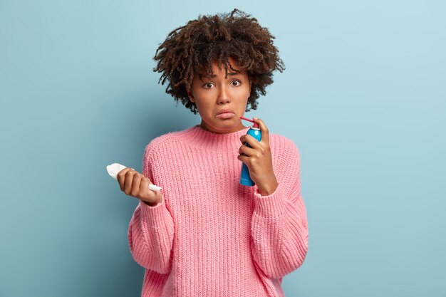 Conceito de pessoas, tratamento e doença. Mulher afro-americana descontente cura dor de garganta com spray, sofre de tosse e bronquite, segura um lenço branco, franze o rosto e usa um macacão rosa