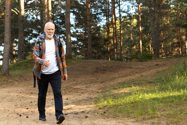 Conceito de saúde, atividade, bem-estar, idade e pessoas. Homem europeu barbudo ativo e autodeterminado na casa dos sessenta anos caminhando rápido durante uma caminhada em uma floresta de montanha, com olhar focado e confiante