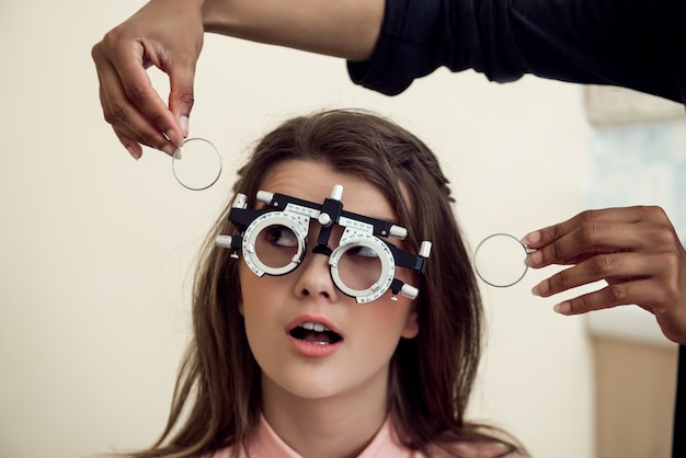 Foto grátis conceito de saúde e olhos. retrato de jovem europeia curiosa e divertida, sentada na cadeira, enquanto um especialista em cuidados oculares testando a vista com um foróptero, perguntando se ela pode ver claramente