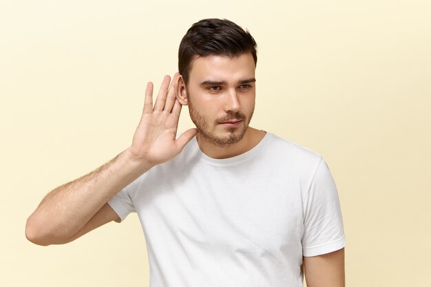 Conceito de segredo e conspiração. Imagem isolada de cara jovem e bonito intrometido com a barba por fazer, segurando a mão na orelha, olhando curioso, bisbilhotando, tentando ouvir uma conversa particular