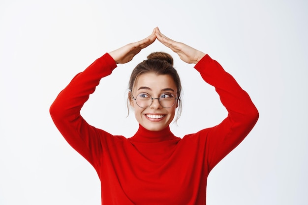 Conceito de seguro e imobiliário Jovem mulher sorridente em óculos fazendo telhado mostrando o telhado de casa com as mãos acima da cabeça, olhando de lado para o fundo branco do espaço de cópia