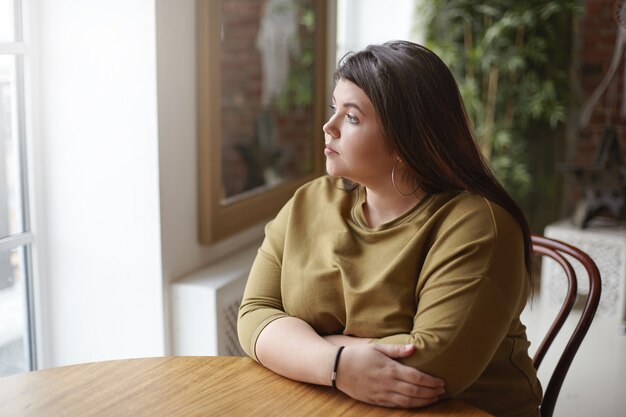 Conceito de solidão. Jovem morena plus size mulher com cabelos pretos sentada à mesa do café, sentindo-se sozinha, passando um tempo sozinha, esperando seu almoço, olhando pela janela com uma expressão triste e pensativa