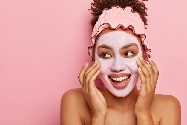 Foto grátis conceito de spa e cuidados com a pele. mulher afro-americana satisfeita aplica máscara nutritiva de argila no rosto, tem expressão alegre, olha para o lado esquerdo, toca a bochecha, luta com problema de pele seca, tem corpo em topless