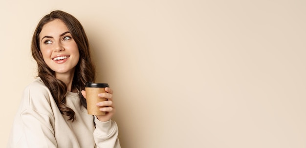 Conceito de takeaway e café linda mulher feminina sorrindo segurando uma xícara de café posando contra beig