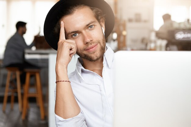 Conceito de tecnologia e comunicação. Jovem elegante com barba e chapéu preto, ouvindo música em fones de ouvido brancos, sentado em frente a um laptop genérico, apoiado no cotovelo e sorrindo