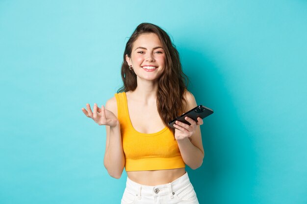 Conceito de tecnologia e estilo de vida. Linda mulher caucasiana conversando no telefone, segurando o smartphone e sorrindo para a câmera, de fundo azul.