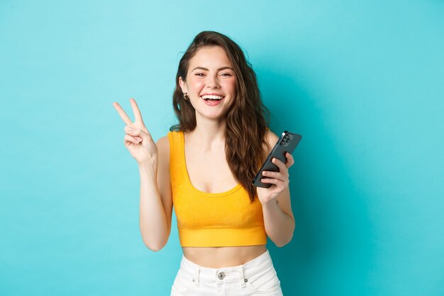 Conceito de tecnologia e estilo de vida. Mulher atraente feliz rindo, mostrando o sinal V e usando o smartphone, conversando no celular, em pé sobre um fundo azul