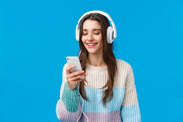 Conceito de tecnologia, geração do milênio e estilo de vida. Despreocupada aluna morena bonita colocar fones de ouvido, plug-in smartphone escolhendo música e sorrindo, fundo azul de pé fazer lista de reprodução para estudo.