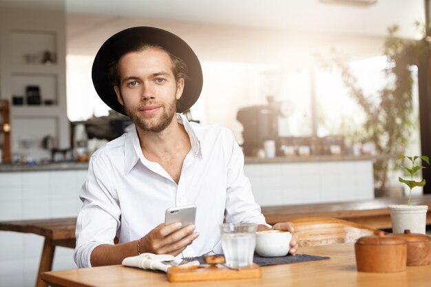 Conceito de tecnologia humana e moderna. Retrato de um jovem estudante caucasiano bonito de chapéu preto e camisa branca, navegando na internet em seu telefone inteligente, desfrutando de conexão sem fio gratuita durante o almoço