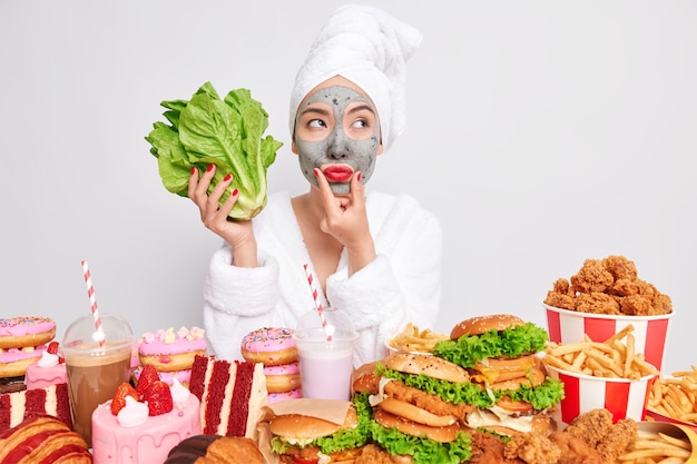 Conceito de tentação de nutrição adequada de estilo de vida saudável. Mulher asiática pensativa segurando salada de alface verde e escolhe entre comida saudável e não saudável